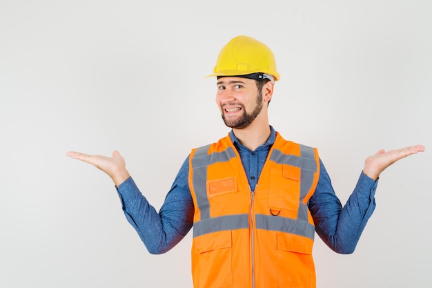 Joven constructor haciendo gestos de escalas en camisa, chaleco, casco y mirando alegre, vista frontal.