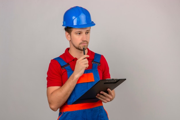 Joven constructor hombre en uniforme de construcción y casco de seguridad de pie con portapapeles en la mano y tocando la barbilla con lápiz con cara seria sobre pared blanca aislada