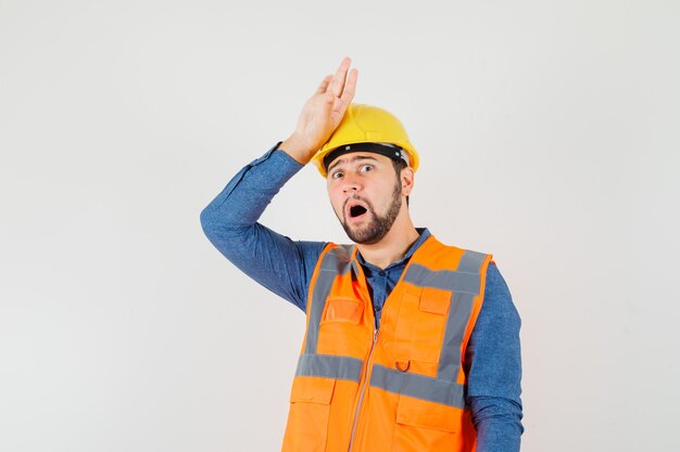 Joven constructor de la mano en la cabeza en camisa, chaleco, casco y mirando nostálgico, vista frontal.