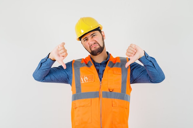 Joven constructor mostrando doble pulgar hacia abajo en camisa, chaleco, casco y mirando insatisfecho. vista frontal.