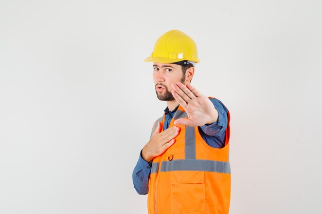 Joven constructor mostrando gesto de parada en camisa, chaleco, casco y mirando resuelto. vista frontal.