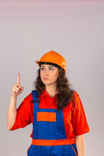 Joven constructor mujer en construcción uniforme y casco de seguridad de pie con el dedo hacia arriba advertencia de peligro sobre la pared blanca aislada