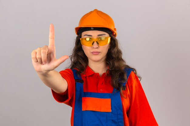 Joven constructor mujer en construcción uniforme y casco de seguridad de pie con el dedo hacia arriba advertencia de peligro sobre la pared blanca aislada