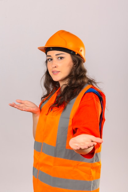 Joven constructor mujer en construcción uniforme y casco de seguridad sonriendo haciendo un gesto de bienvenida sobre la pared blanca aislada