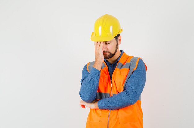 Joven constructor de pie en pose de pensamiento en camisa, chaleco, casco y mirando cansado, vista frontal.