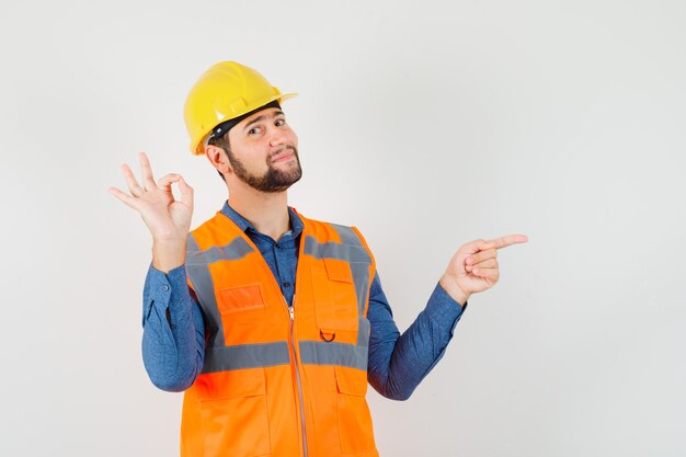 Joven constructor que muestra el signo de ok, apuntando hacia el lado en camisa, chaleco, casco y mirando alegre. vista frontal.