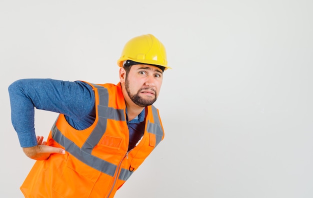 Joven constructor que sufre de dolor de espalda en camisa, chaleco, casco y mirando fatigado, vista frontal.