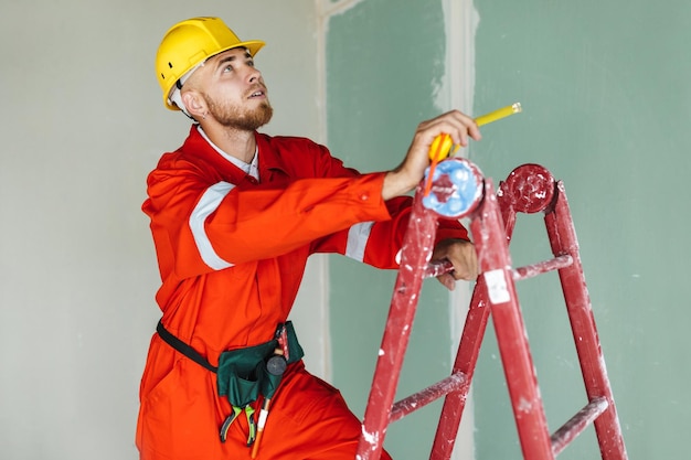 Joven constructor con ropa de trabajo naranja y casco amarillo parado en la escalera con cinta métrica en la mano mirando cuidadosamente el techo en un piso nuevo