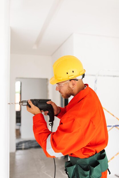 Joven constructor en ropa de trabajo naranja y casco amarillo con taladro eléctrico para hacer un agujero en la pared