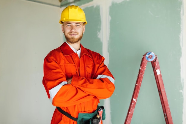 Joven constructor sonriente con ropa de trabajo naranja y casco amarillo mirando felizmente a la cámara con una escalera en el fondo