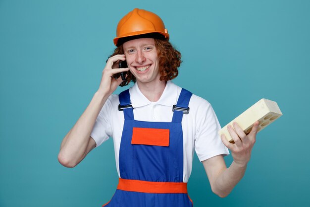 Un joven constructor sonriente con uniforme sosteniendo ladrillos y hablando por teléfono aislado de fondo azul