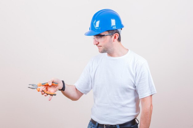 Joven constructor sosteniendo alicates en camiseta, jeans, casco y mirando confiado