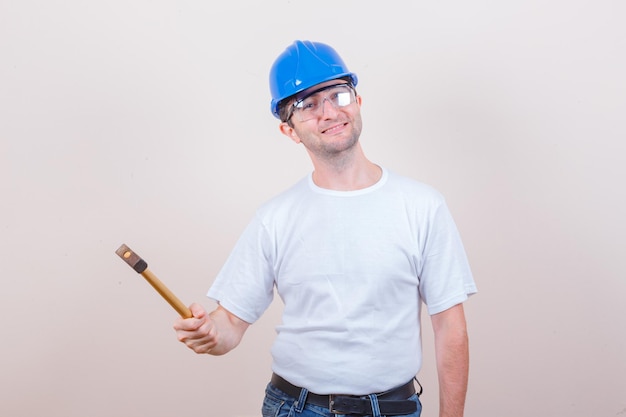 Joven constructor sosteniendo un martillo en camiseta, jeans, casco y mirando alegre