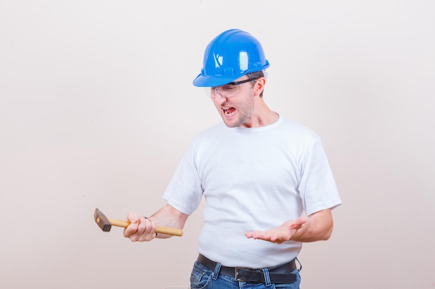 Joven constructor sosteniendo un martillo mientras grita en camiseta, jeans y casco