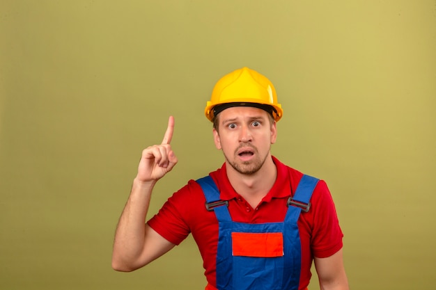 Joven constructor en uniforme de construcción y casco de seguridad con cara sorprendida apuntando el dedo hacia arriba sobre la pared azul aislada