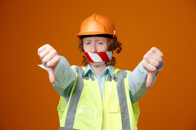 Joven constructor en uniforme de construcción y casco de seguridad con cinta adhesiva en la boca mirando a la cámara insatisfecho mostrando los pulgares hacia abajo de pie sobre fondo naranja