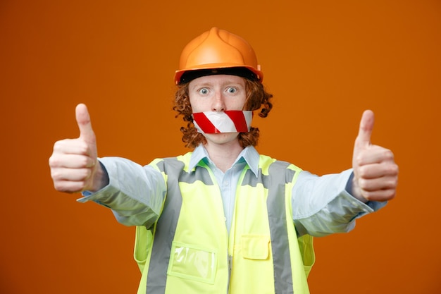 Joven constructor en uniforme de construcción y casco de seguridad con cinta adhesiva en la boca mirando a la cámara sorprendido mostrando los pulgares hacia arriba de pie sobre fondo naranja