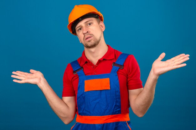 Joven constructor en uniforme de construcción y casco de seguridad despistado y expresión confusa con brazos y manos planteó el concepto de duda sobre la pared azul aislada