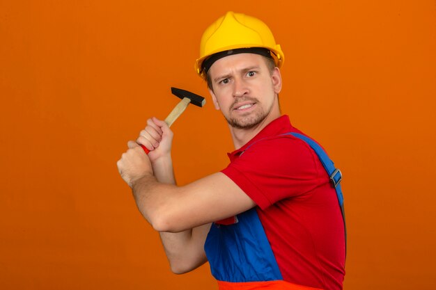 Joven constructor en uniforme de construcción y casco de seguridad con expresión enojada que amenaza con golpear con un martillo sobre una pared naranja aislada