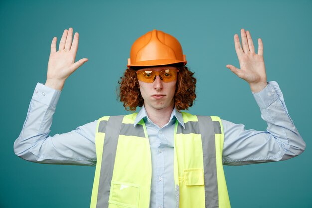 Joven constructor en uniforme de construcción y casco de seguridad con gafas de seguridad mirando a la cámara con cara seria levantando los brazos de pie sobre fondo azul.