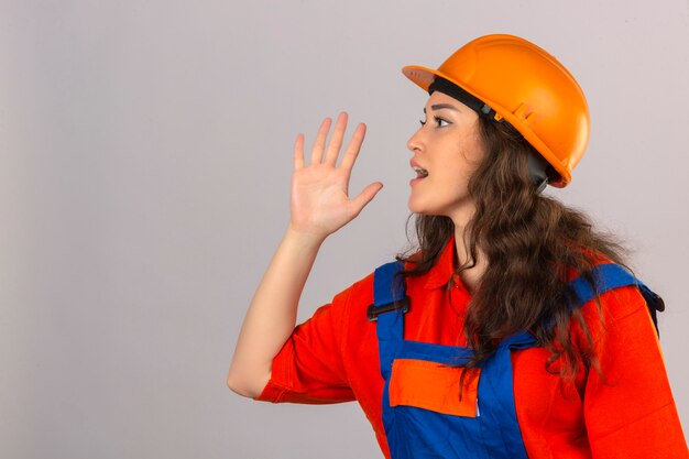 Joven constructor en uniforme de construcción y casco de seguridad gritando a un lado con la mano en la boca sobre la pared blanca aislada