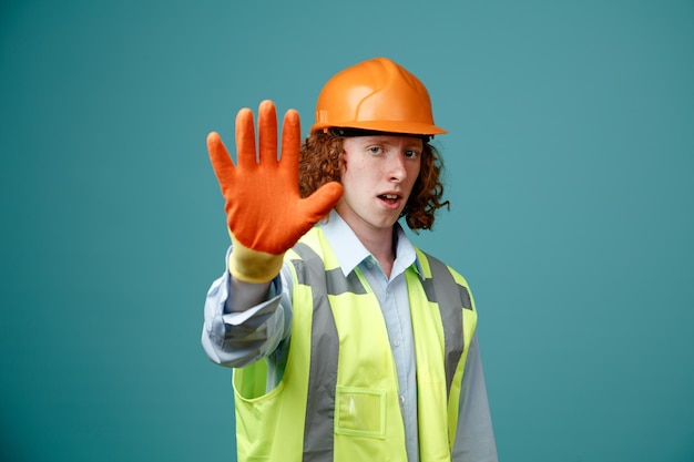 Joven constructor con uniforme de construcción y casco de seguridad con guantes de goma mirando a la cámara con cara seria haciendo un gesto de parada mostrando la palma abierta de pie sobre fondo azul