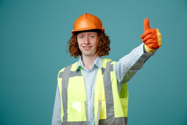 Joven constructor en uniforme de construcción y casco de seguridad con guantes de goma mostrando el pulgar hacia arriba sonriendo feliz y positivo de pie sobre fondo azul.