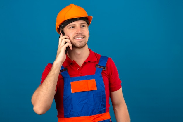 Joven constructor en uniforme de construcción y casco de seguridad hablando por teléfono móvil con cara feliz sonriendo sobre pared azul aislado