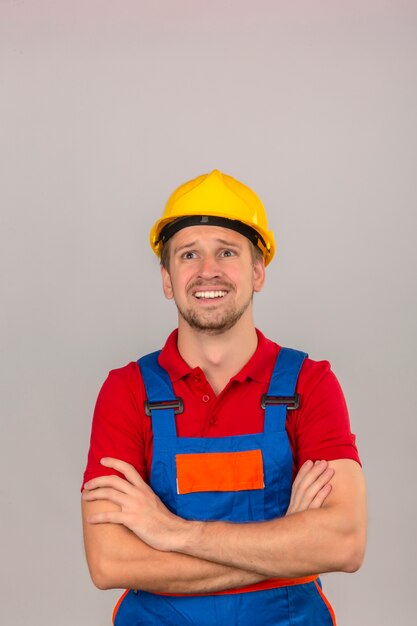 Joven constructor en uniforme de construcción y casco de seguridad mirando hacia arriba con cara de miedo sobre pared blanca aislada