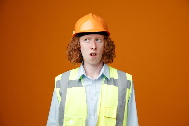 Joven constructor en uniforme de construcción y casco de seguridad mirando hacia arriba pensando desconcertado de pie sobre fondo naranja