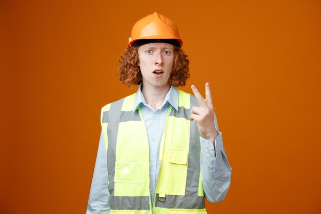 Joven constructor en uniforme de construcción y casco de seguridad mirando a la cámara confundido mostrando el número dos con los dedos sobre fondo naranja