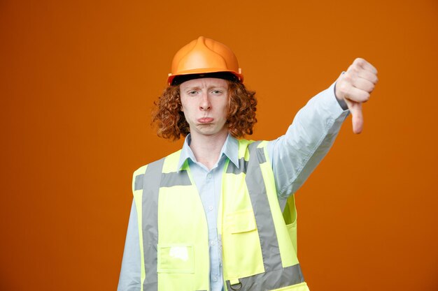 Joven constructor en uniforme de construcción y casco de seguridad mirando a la cámara insatisfecho haciendo la boca torcida mostrando el pulgar hacia abajo de pie sobre fondo naranja