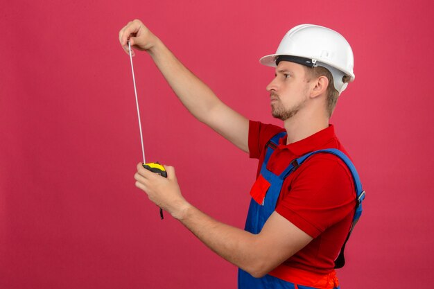Joven constructor en uniforme de construcción y casco de seguridad mirando cinta métrica en sus manos mirada pensativa sobre pared rosa aislado