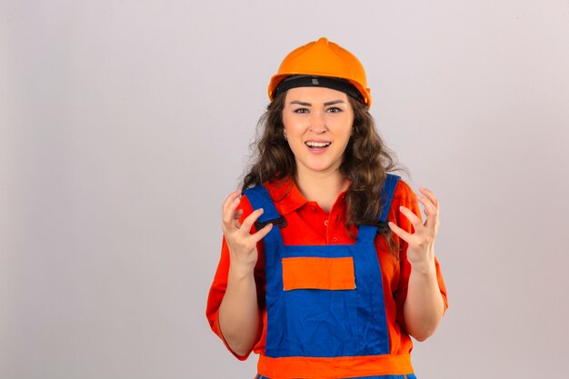 Joven constructor en uniforme de construcción y casco de seguridad mirando gritos frustrados con expresión agresiva y los brazos levantados sobre la pared blanca aislada