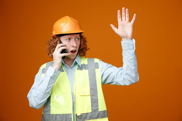 Joven constructor con uniforme de construcción y casco de seguridad mirando a un lado confundido levantando el brazo con disgusto mientras habla por teléfono móvil de pie sobre un fondo naranja