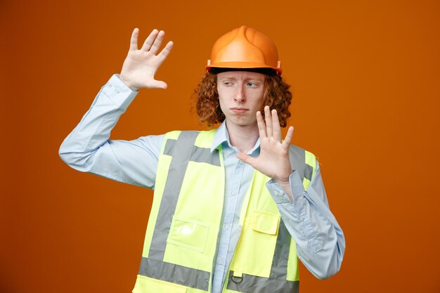 Joven constructor en uniforme de construcción y casco de seguridad mirando a un lado haciendo gesto de defensa con las manos sobre fondo naranja