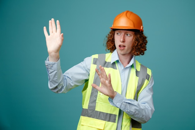 Joven constructor con uniforme de construcción y casco de seguridad mirando a un lado preocupado haciendo un gesto de defensa con las manos sobre fondo azul