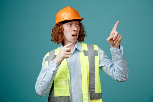 Joven constructor en uniforme de construcción y casco de seguridad mirando a un lado sorprendido señalando con los dedos índices hacia un lado de pie sobre fondo azul