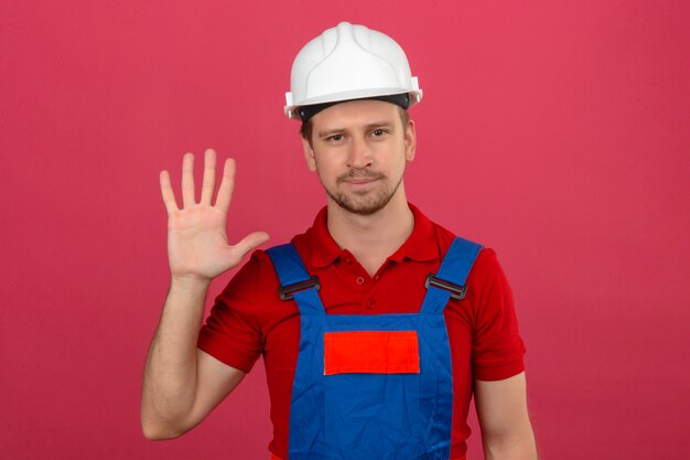 Joven constructor en uniforme de construcción y casco de seguridad mostrando y apuntando hacia arriba con los dedos número cinco con una sonrisa en la cara sobre la pared rosa aislada