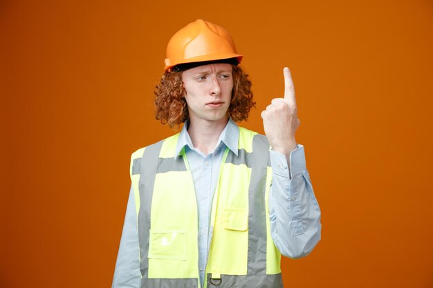 Joven constructor en uniforme de construcción y casco de seguridad mostrando el dedo índice mirándolo con cara seria de pie sobre fondo naranja