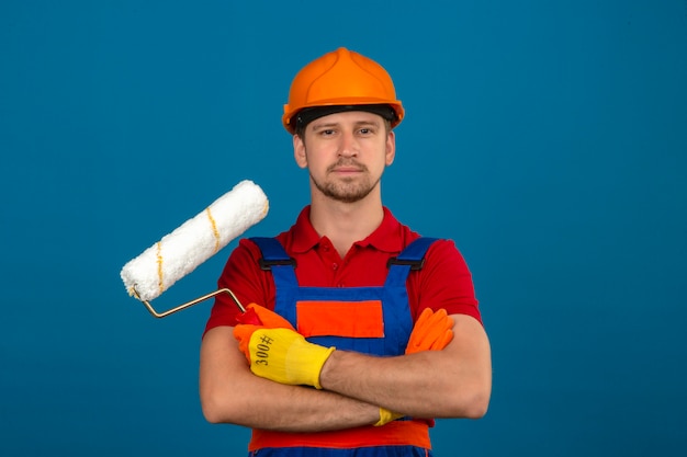 Foto gratuita joven constructor en uniforme de construcción y casco de seguridad de pie con los brazos cruzados con rodillo de pintura mirando confiado sobre la pared azul aislada