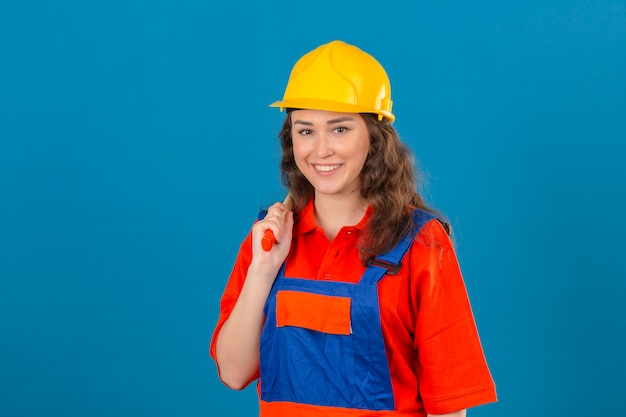 Joven constructor en uniforme de construcción y casco de seguridad de pie con un martillo en el hombro sonriente y feliz sobre la pared azul aislada