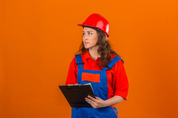 Joven constructor en uniforme de construcción y casco de seguridad de pie con portapapeles mirando a otro lado sobre la pared naranja aislada