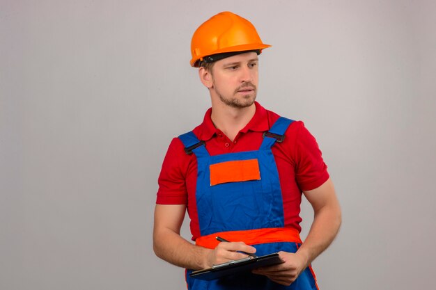 Joven constructor en uniforme de construcción y casco de seguridad con portapapeles y mirando hacia el lado con cara seria sobre pared blanca aislada