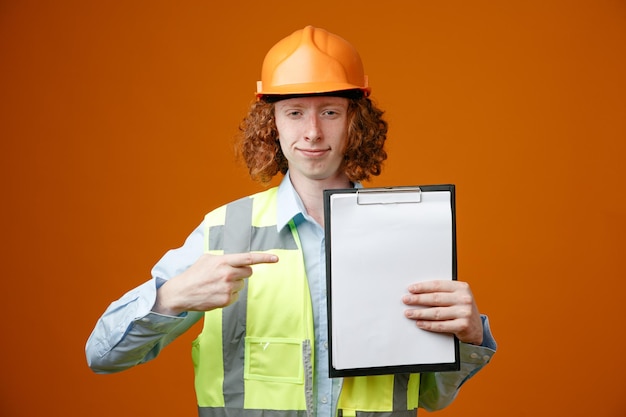 Joven constructor en uniforme de construcción y casco de seguridad que muestra el portapapeles apuntando con el dedo índice mirando confiado de pie sobre fondo naranja