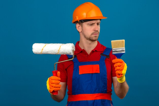 Foto gratuita joven constructor en uniforme de construcción y casco de seguridad con rodillo de pintura y pincel mirándolo con expresión escéptica de la cara sobre la pared azul aislada