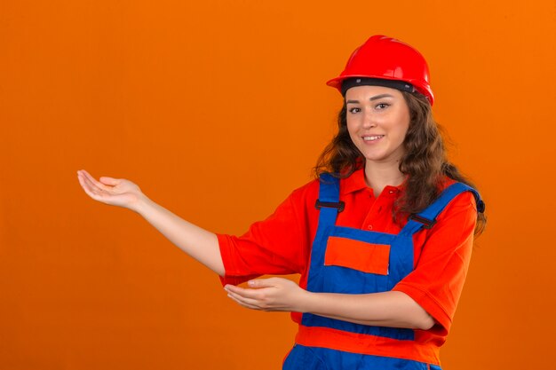 Joven constructor en uniforme de construcción y casco de seguridad sonriendo alegre presentando y señalando con las palmas de las manos mirando a la cámara sobre la pared naranja aislada