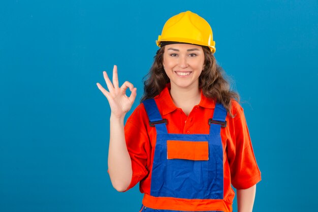 Joven constructor en uniforme de construcción y casco de seguridad sonriendo alegremente haciendo bien firmar sobre pared azul aislado