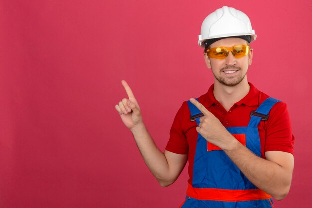 Joven constructor en uniforme de construcción y casco de seguridad sonriendo y mirando a la cámara apuntando con las dos manos y los dedos a un lado sobre la pared rosa aislada con espacio de copia