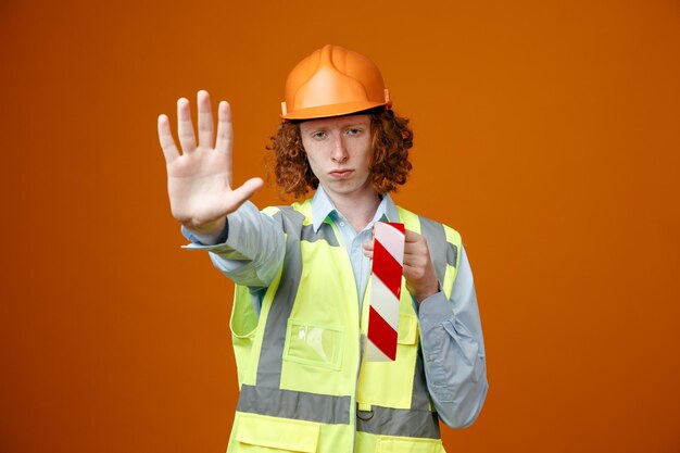 Joven constructor en uniforme de construcción y casco de seguridad sosteniendo cinta adhesiva mirando a la cámara con cara seria haciendo un gesto de parada con la mano de pie sobre fondo naranja